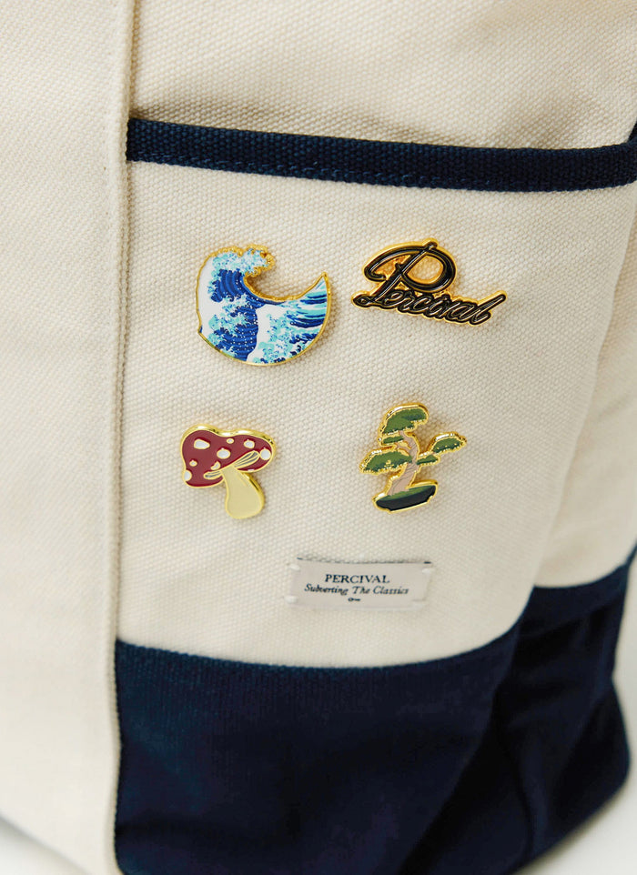 A collection of Percival's colourful metal pin badges, including the script logo, pinned onto a cream canvas bag.