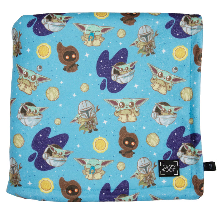 The Sassy Woof STAR WARS™ The Mandalorian™ pet blanket features a blue microfiber top with cartoon Baby Yoda, Mandalorian, Jawas, purple stars, and yellow coins. Made from 100% polyester.