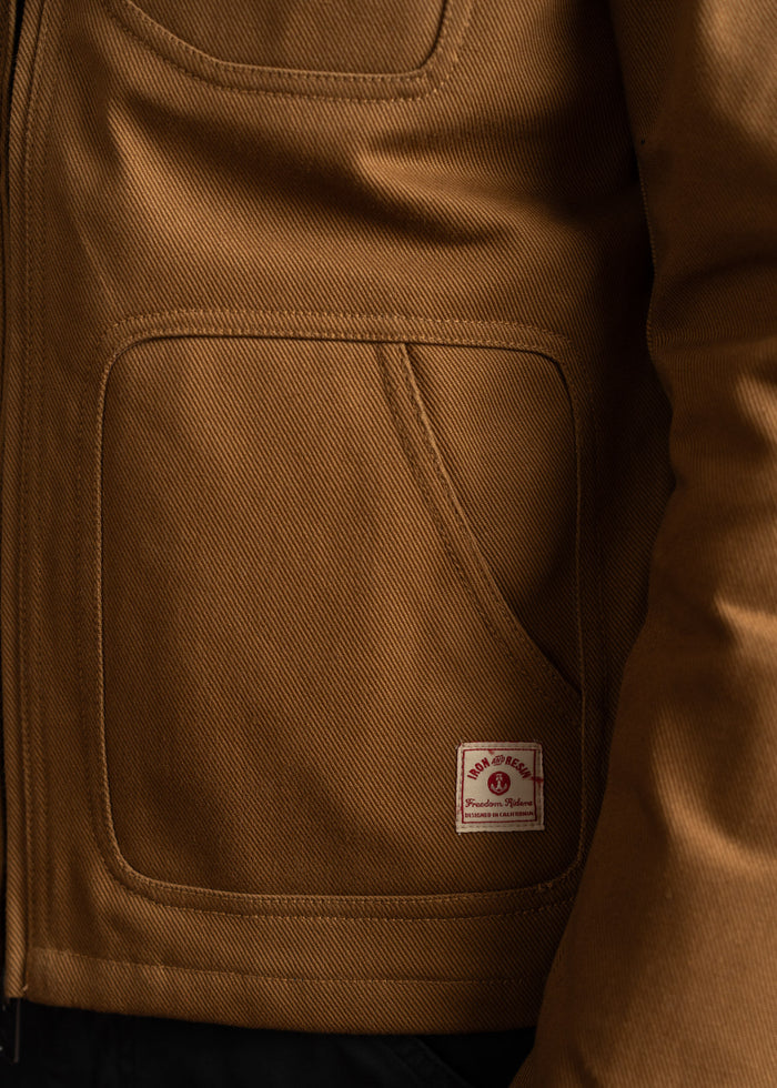 Close-up of a brown jacket pocket with a visible "Iron and Resin" label, highlighting the rugged fabric texture and stitching.