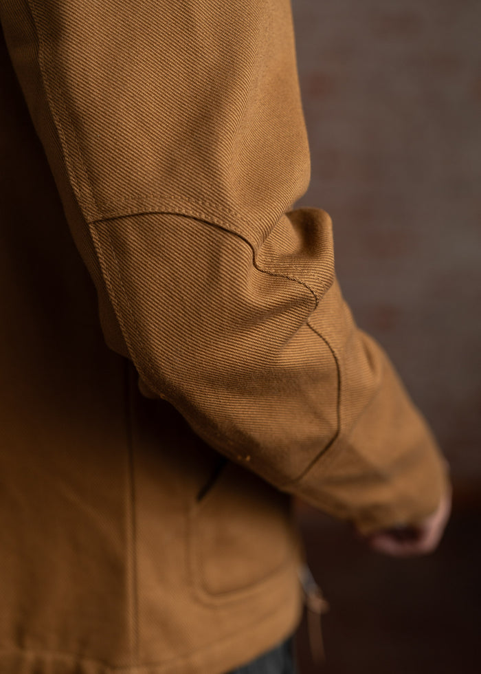 Close-up of a brown jacket sleeve with visible stitching and texture, set against a blurred background, highlighting the fabric detail.