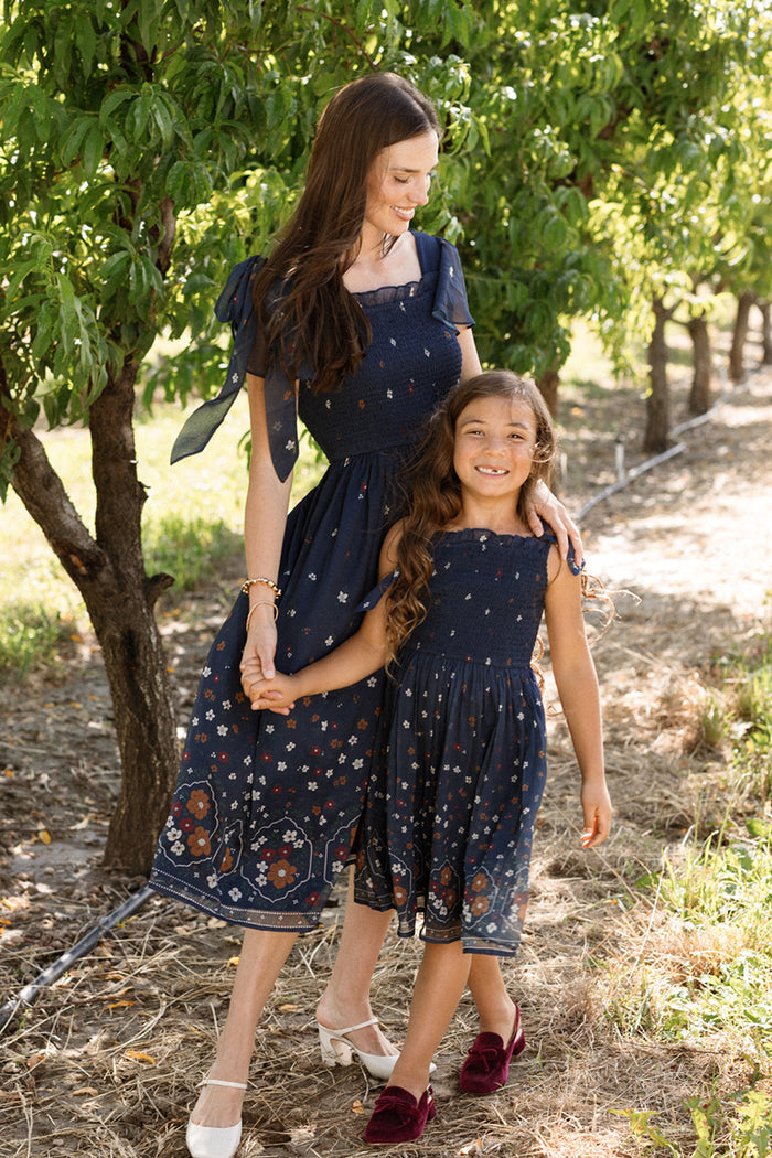 A woman and a girl, both wearing the Ivy City Co Rayna Dress in Midnight Cascading Floral—with matching navy blue chiffon fabric, floral patterns, and smocked bodices—hold hands and smile as they stand outdoors among green trees on a sunny day.
