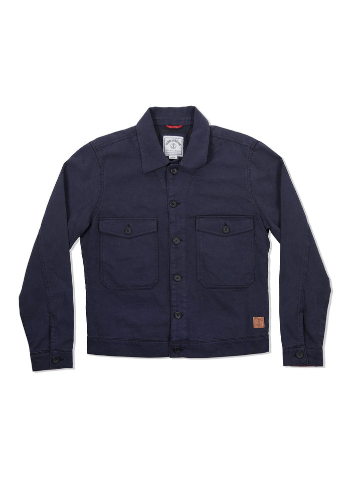 Dark blue denim jacket with buttons, two chest pockets, and a small brown patch on the lower right. Simple, classic design.