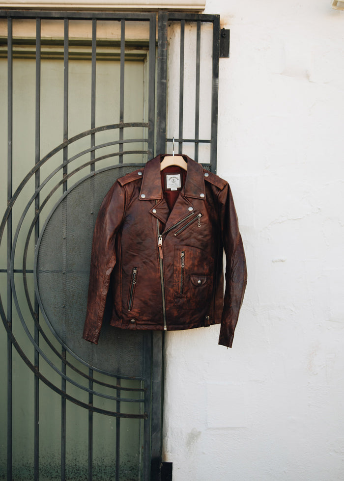Brown leather jacket hangs on a metal gate against a white wall, showcasing its vintage style and detailed zippers.