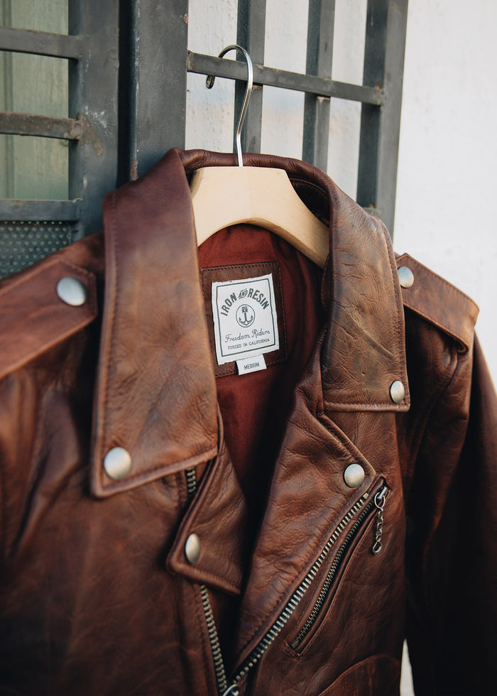 Brown leather jacket with silver buttons and zippers hangs on a wooden hanger. Iron and Resin label visible inside the collar.