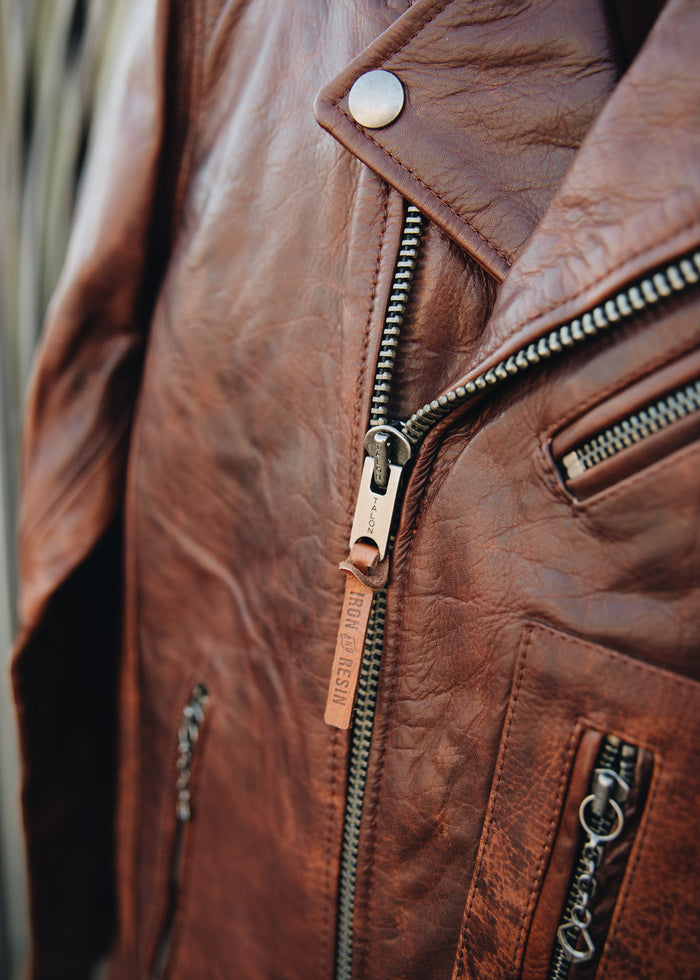 Close-up of a brown leather jacket with a prominent zipper and metal snap, highlighting its texture and craftsmanship.
