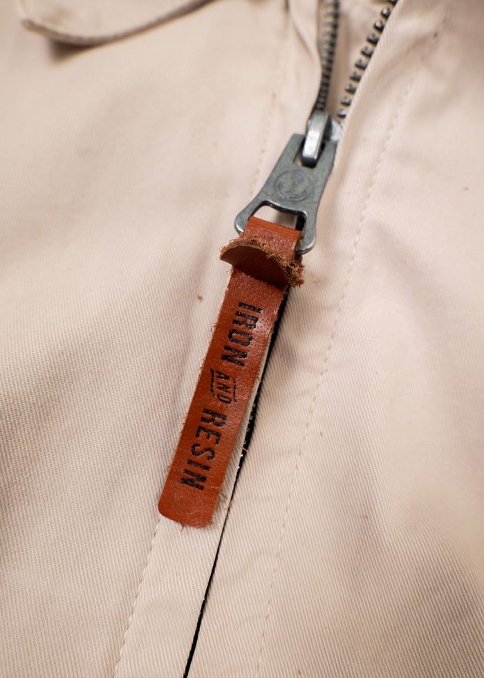 Close-up of a beige jacket zipper with a brown leather pull tab labeled "Iron and Resin," highlighting the brand detail.