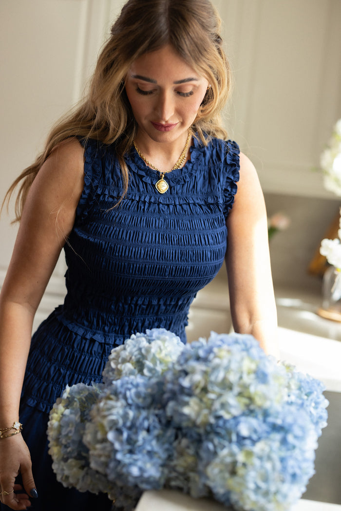 A woman wearing the Simone Dress in Navy by Ivy City Co stands indoors, looking down at a bouquet of light blue hydrangeas. She has wavy, light brown hair and accessorizes with a gold necklace.