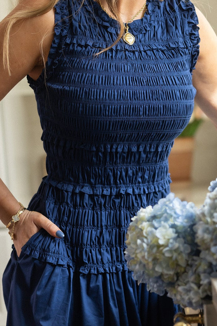 A woman in Ivy City Co’s Simone Dress in Navy—a maternity-friendly, sleeveless cotton dress with a smocked bodice and pockets—accessorizes with a gold necklace and bracelet, with a blue and white hydrangea bouquet in the foreground.