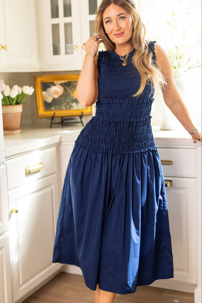 A woman with long blonde hair wears the maternity-friendly Simone Dress in Navy by Ivy City Co while standing in a bright kitchen with white cabinets, flowers, and a small framed painting on the counter.