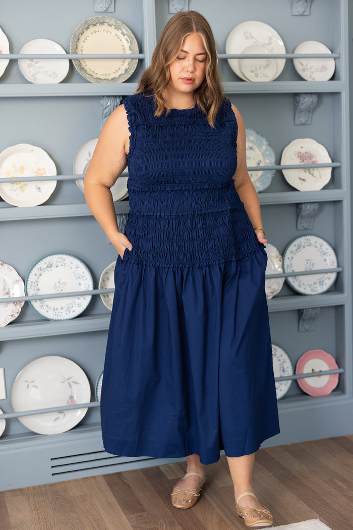 A woman stands indoors in front of shelves with decorative plates, wearing the Ivy City Co Simone Dress in Navy, a sleeveless cotton style with a smocked bodice and pockets. She has light brown hair, transparent sandals, and the dress is maternity friendly.