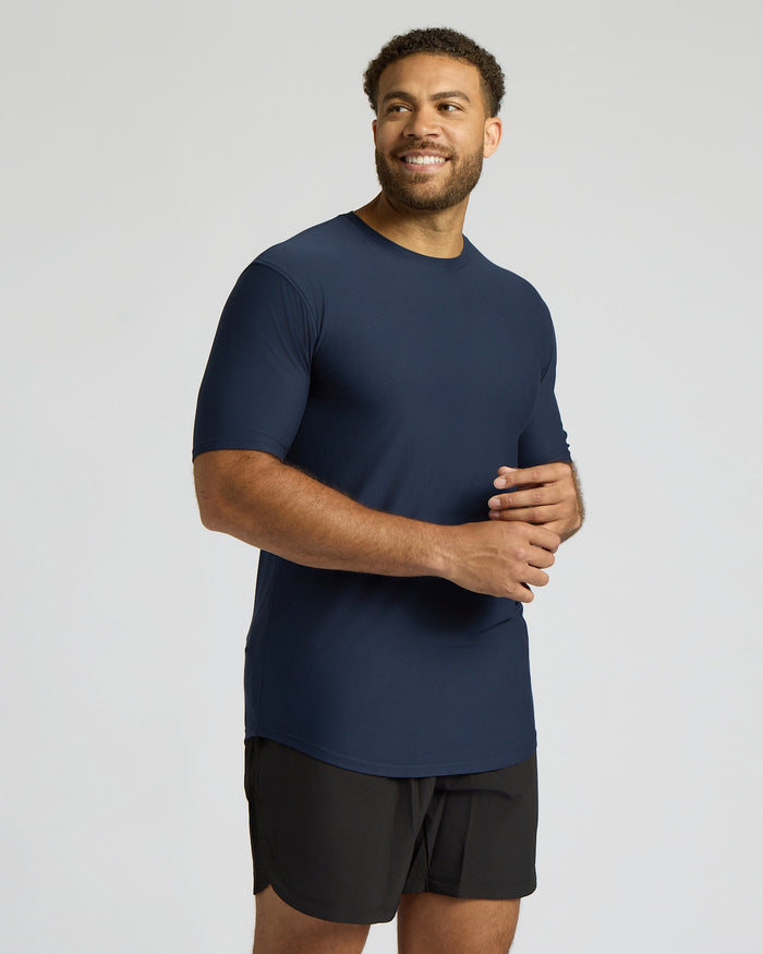 Man wearing a navy blue Tall Active Curved Hem Crew 3-Pack shirt and black shorts, standing smiling against a light gray background in a relaxed pose.