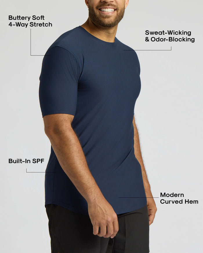 A man wearing a navy blue short-sleeve athletic t-shirt, shown from the chest down against a light gray background, with text callouts highlighting features like 4-way stretch, sweat-wicking, built-in SPF, and modern curved hem.