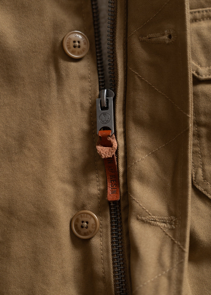 Close-up of a brown jacket's zipper and buttons, featuring a leather pull tab with embossed text, highlighting craftsmanship.