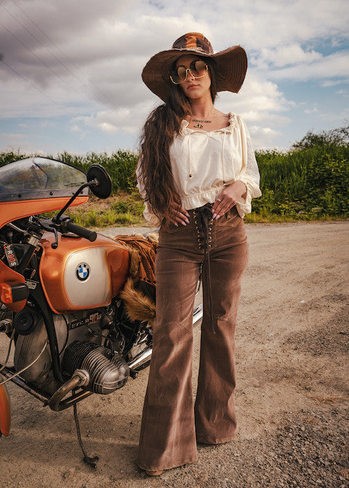 Woman in vintage attire standing next to a BMW motorcycle on a road with a scenic background wearing Soul Sister since 1969
