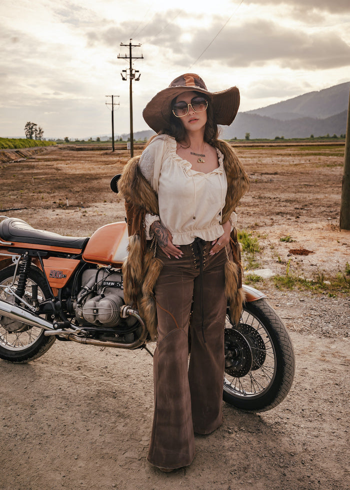 Person in vintage attire standing next to a motorcycle in an open field with mountains in the background wearing Soul Sister since 1969