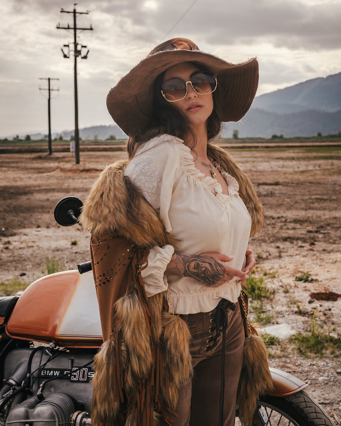 Woman in rustic attire with fur vest and wide-brimmed hat standing next to a motorcycle in an open field wearing Soul Sister since 1969.