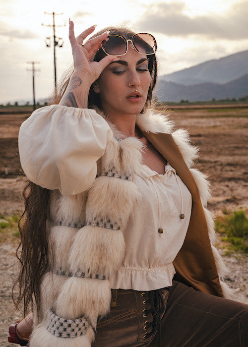 Woman in a stylish outfit with fur accents sitting outdoors in a field with mountains in the background wearing Soul Sister since 1969.