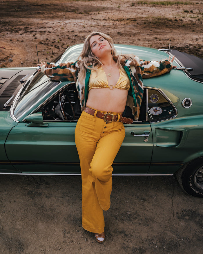 Woman in a gold bikini top and yellow pants leaning against a green vintage car in a desert setting wearing Soul Sister since 1969.