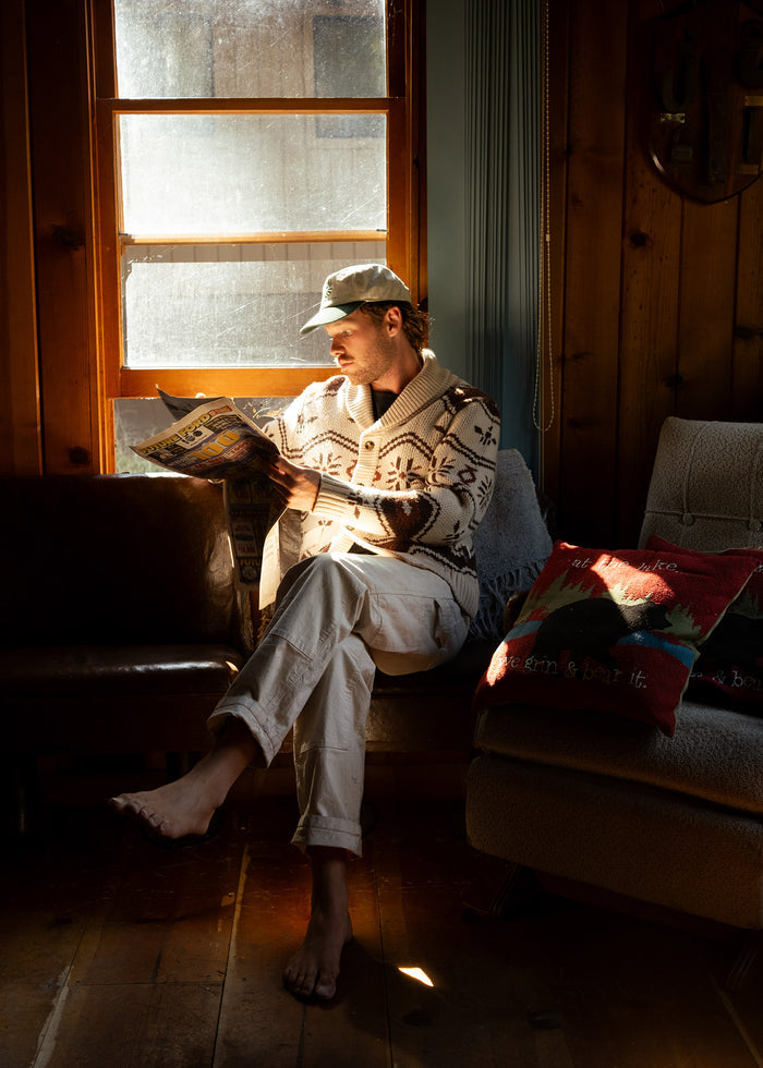 Man in a cozy sweater and cap reads a magazine by a sunlit window, sitting barefoot on a couch with colorful pillows.
